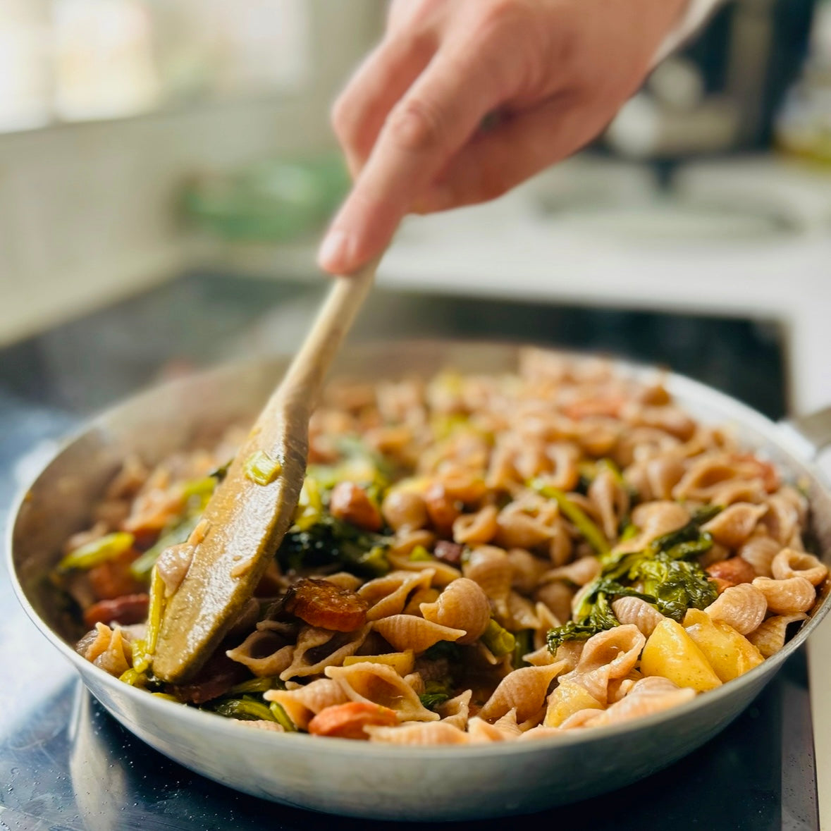 Person stirring wholegrain organic pasta with vegetables in a pan using a wooden spoon.
